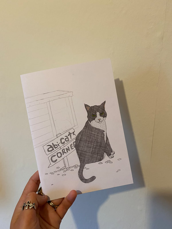 Photo of a hand wearing silver rings holding a line drawing of a black and white cat sat in front of a wooden hutch and a sign that reads 'Abi-Cat Corner'. The drawing uses fine lines and various mark making to build up texture and tone alongside pro marker pens to highlight colour.