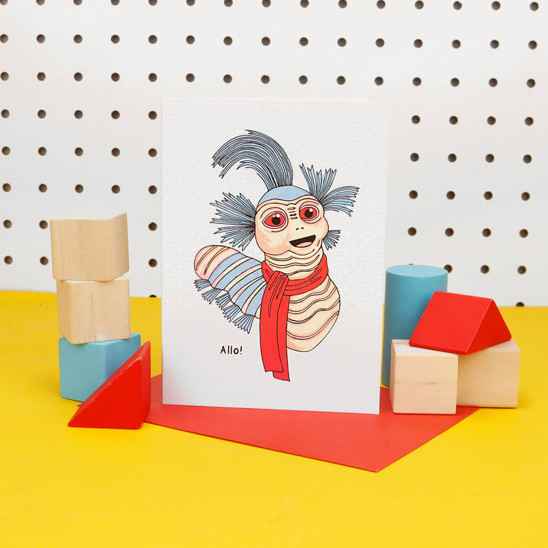 Labyrinth worm greetings card stood up on a red envelope with children's red, blue and natural wood building blocks on either side.