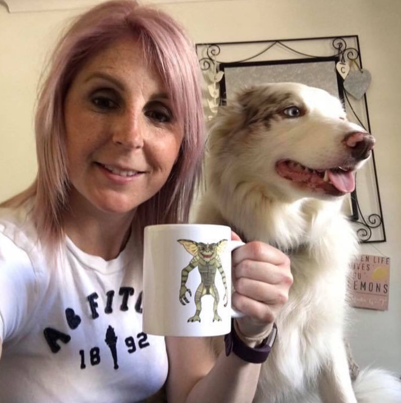 Smiling blond woman sat beside her white and sandy coloured long haired blue eyed dog holding a cup with the bad gremlin Stripe on it.