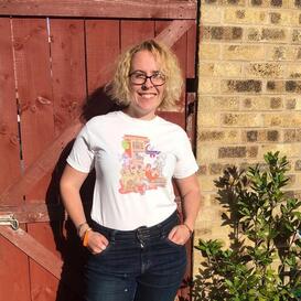 Rebecca (blond curly hair and glasses) smiling in the sunshine wearing one of her Ghostbusters T-shirts and jeans.